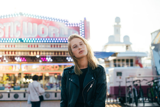 Beautiful fashion model with blonde hair, wears black leather jacket and stands in middle of carnvial, festival or fair, in front of attraction rides during summer time holidays or spring break