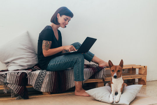 Pretty woman works from home or startup coworking space, sits barefoot on bench and writes code or blog on laptop, her best friend dog puppy lays next to her on pillow