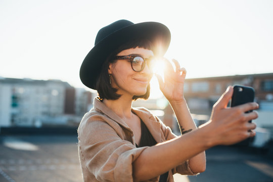 Beautiful and stylish attractive young woman or teenager makes selfie on smartphone using amazing sunset light at rooftop of in open space in big city, smiles and poses like fashion model