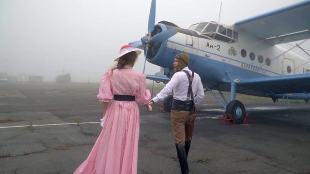 A rotating camera, a male pilot in a helmet, leads a woman in a pink dress and a white hat to a retro airplane