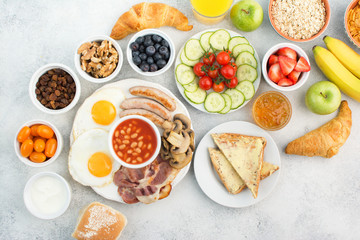 Healthy varied breakfast, fried eggs, sausages, bacon and mushrooms with selection of fruits and vegetables, breads and juice on the grey white table, top view, copy space for text, selective focus