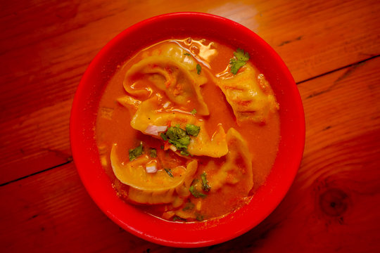 Top View Of Steamed Soup Of Momo Served In A Red Bowl. A Popular Nepalese Food That Is Also Common In Chiana, Bhutan, Tibet And India, In A Wooden Table Background