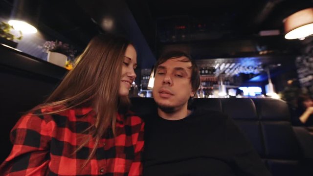 Young attractive couple is having a good time in a cafe: looking into the camera, lady is smiling, man is kissing her on a cheek and speaking to someone behind the camera. Camera is zooming in