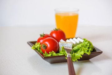 Tomatoes and cheese slices lie on a sheet of fresh salad in a plate. A glass of orange juice stands side by side on the table.