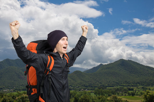 Young man with backpack standing on top of a mountain with his hands up.