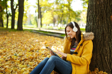 A brown-haired smiling woman in yellow coat and jeans sitting and listening to music under a tree...