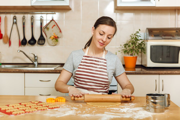 A young beautiful happy woman 30-35 years in apron sitting at a table with dough and rolling pin and going to prepare a Christmas ginger cakes in the light kitchen. Cooking home. Prepare food.