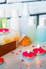 Fototapeta na wymiar Spa accessories. Small lighted candles, many colored soaps of soap, many different shapes of bottles for the bathroom. The bath salt is pink, the body scrub is showered. Light brick and white wooden a