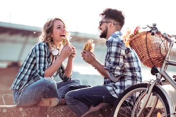 Young man and woman are siting on the curb and eat sandwiches.
