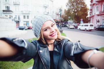 Young woman taking selfie on the street.Closeup selfie-portrait  of student of attractive girl in black leather jacket  with long hairstyle and snow-white smile in city.n black rock style over city
