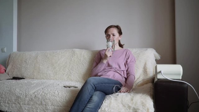 A woman sits on the couch at home and inhales through an inhaler. Inhalation mask on the face.Treatment at home.