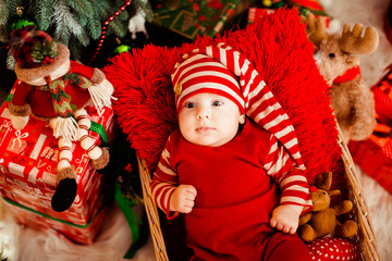 Little boy in funny red suit and long hat lies in the basket before a Christmas tree