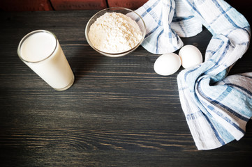 Transparent plate bowl with flour. Whisk for whipping with a blender. A shell of chopped eggs. Milk in a measuring glass. The wall is made of red bricks, the table is made of ebony. Cooking process