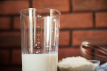 Transparent plate bowl with flour. Whisk for whipping with a blender. A shell of chopped eggs. Milk in a measuring glass. The wall is made of red bricks, the table is made of ebony. Cooking process