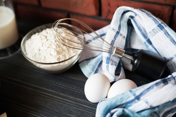 Transparent plate bowl with flour. Whisk for whipping with a blender. A shell of chopped eggs. Milk in a measuring glass. The wall is made of red bricks, the table is made of ebony. Cooking process