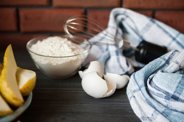 Transparent plate bowl with flour. Whisk for whipping with a blender. A shell of chopped eggs. Pears in the background. The wall is made of red bricks, the table is made of ebony. Cooking process