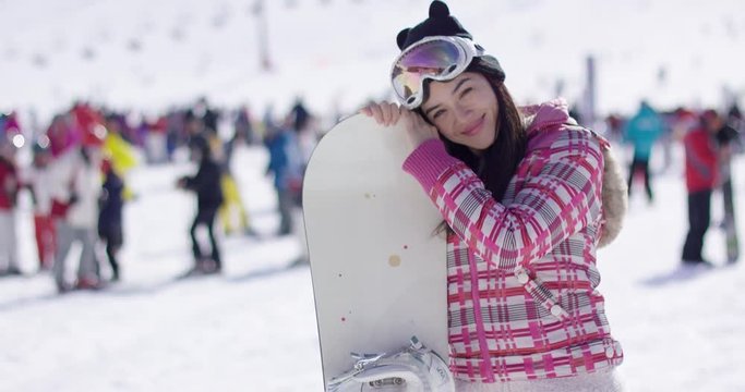 Happy-looking young woman in down parka and goggles standing with snowboard on ski resort, waving her hand and looking at camera.