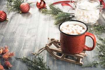 Cup of cocoa on grey wooden background. Red mug of hot chocolate on miniature toy sledge.  Fir-tree branches, tape, Christmas balls