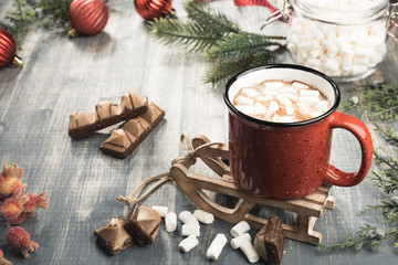 Cup of cocoa on grey wooden background. Red mug of hot chocolate on miniature toy sledge.  Fir-tree branches, tape, Christmas balls, candies