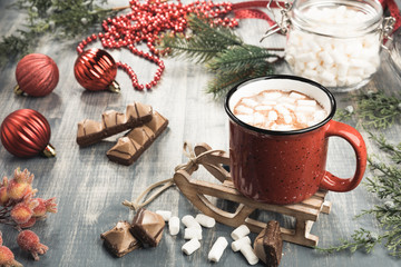 Cup of cocoa on grey wooden background. Red mug of hot chocolate on miniature toy sledge.  Fir-tree branches, tape, Christmas balls, candies