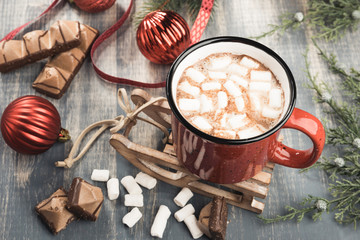 Cup of cocoa on grey wooden background. Red mug of hot chocolate on miniature toy sledge.  Fir-tree branches, tape, Christmas balls, candies