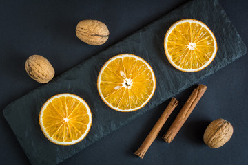 Dry slices of orange fruits, walnuts and cinnamon on a black slate background.