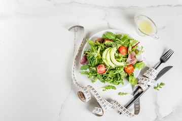 Healthy balanced diet concept, weight loss, calorie counting. Plate with green salad leaves, tomatoes, avocado with yogurt dressing, white table, with fork, knife, measuring tape, top view copy space