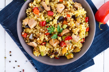 Light healthy dietary salad with couscous, vegetables (zucchini, eggplant, carrots, sweet peppers, onions), chicken pieces on a white wooden background.