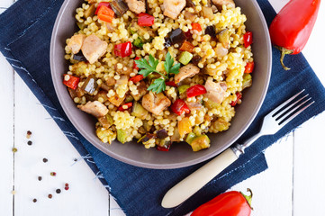 Light healthy dietary salad with couscous, vegetables (zucchini, eggplant, carrots, sweet peppers, onions), chicken pieces on a white wooden background.