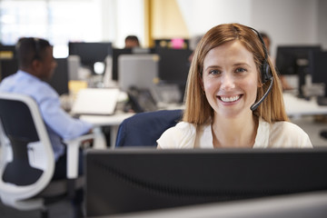 Young woman working at computer with headset in busy office - obrazy, fototapety, plakaty