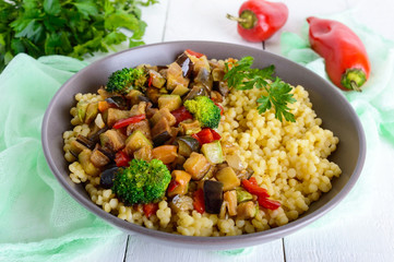 Warm vegan salad with couscous and fried vegetables (eggplant, zucchini, onion, sweet pepper, broccoli) in a ceramic bowl on a white wooden background. Proper nutrition.