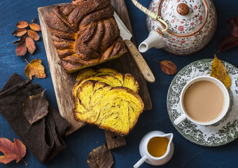 Cut the pumpkin brioche cinnamon on rustic wooden chopping board, tea with milk and a teapot on a blue background, top view. Delicious seasonal pastries