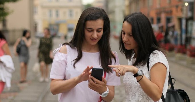 Two pretty girls using the cell phone. Both are happy ang trying to download the app. Outdoors.