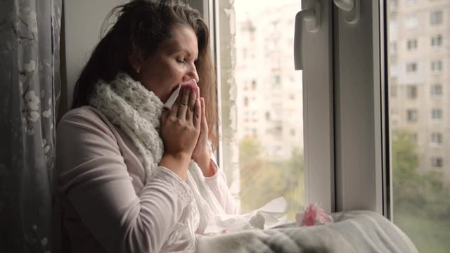 A cold, chilled woman sneezes. Girl blowing his nose while sitting at the window.