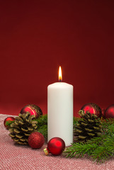 white candle with red christmas baubles and fir cones on red background