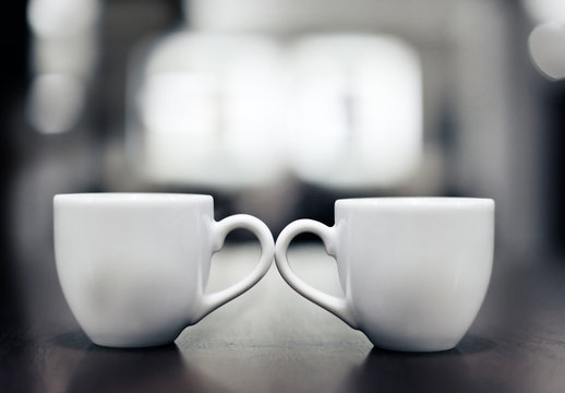 two white coffee cups standing side by side and touching their ears, in the background nice blurry