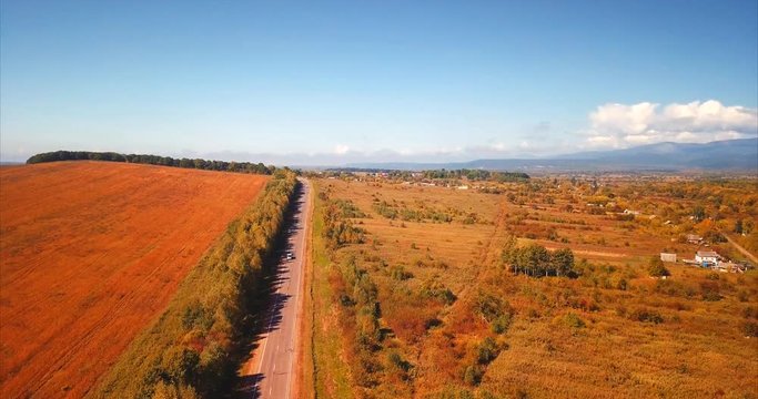 Flying above the highway with driving cars, stubbled field is on the left, and coutryside is on the rigth with mountains on the background. Bright blue sky. September. Russia. Aerial