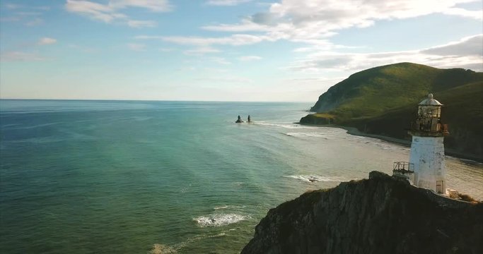 Slowly backwards flying near rocky cliff with lighthouse Brinera on it. It's an octahedral tower in the shape of truncated pyramid with lantern. Amazing seashore is on the background. Russia. Aerial