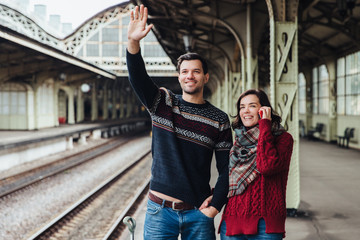 Lovely couple being on platform of railway station, wave someone as look into distance, meet their relatives or friends. Young married woman and man say goodbye someone. Farewell concept.