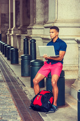 African American College Student traveling, studying in New York, wearing blue short sleeve shirt, red shorts, with backpack on ground, sitting on pillar on street, working on laptop computer..
