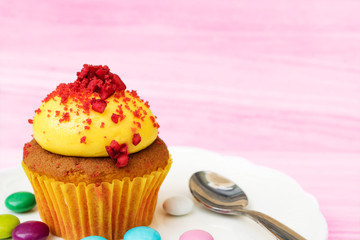 Mini soft cake or cupcake with yellow whipped cream on top with mash cherry, put on the ceramic plate with   colorful chocolate tablets on the pink wooden table background