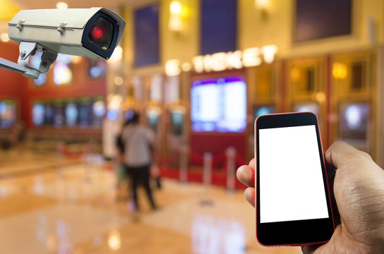 hand holding mobile phone with blank screen and CCTV, security indoor camera system operating at movie theater, internet, surveillance security and safety technology concept