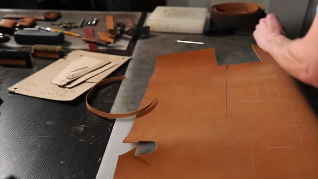 Skinner is cutting out a detail of bag from treated animals skin. There are many tools on his table, master is working on an exclusive leather accessories.
