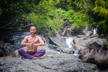An attractive young man sits in a lotus pose and meditates against a background of stones and a waterfall.
