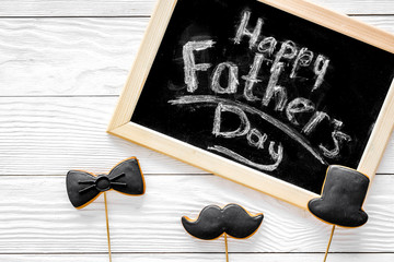 Words happy Father's day written on blackboard. Black tie, mustache and hat cookies. White wooden background top view