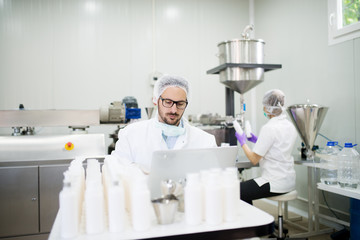 Serious scientist working on a laptop while his assistant doing a production of liquid soap in the...