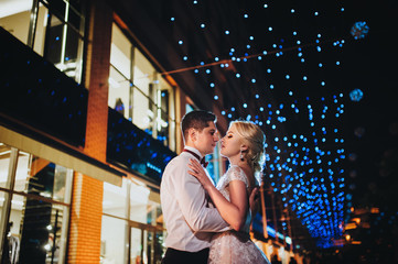 A pair of loving newlyweds gently embrace on the evening city background.