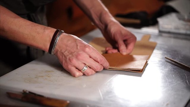Artisan is connecting details of leather luxury cover for notepads. He is putting glued parts to each other and aligning on a table, close-up