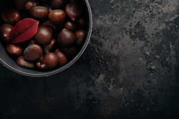 Chestnuts on dark black rustic background. Pile of fresh chestnuts ready to roast in cast iron pan over black antique background. Top view, copy space.