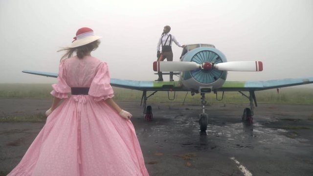 Slow motion shooting as a woman in a pink dress approaches a man in a pilot's suit of 60s that stands on a vintage plane and looks into the distance